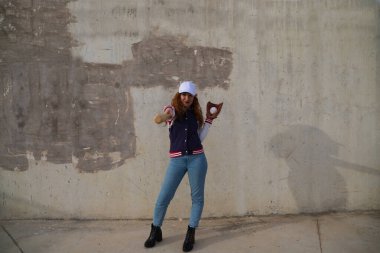 Genç ve güzel kızıl saçlı beysbol şapkalı, ceketli, beysbol sopalı kameralı bir kadın. Gri beton arka planda. Spor ve eğlence konsepti