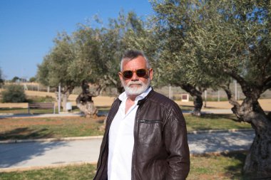 Sakallı ve gri saçlı yakışıklı olgun bir adam parkta yürüyor. Adam bej pantolon, beyaz gömlek ve deri ceket giyiyor. Yaşlılık kavramı