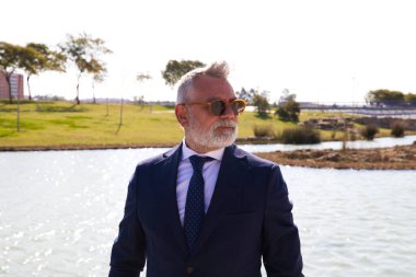 Yakışıklı olgun bir adam, yönetici, gri saç, sakal, güneş gözlüğü, ceket ve kravat, parkta fotoğraf için suni bir göl ile poz veriyor. Olgun adam, büyükbaba, yönetici kavramı.