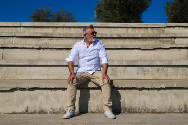 Sakallı ve gri saçlı yakışıklı olgun bir adam açık hava konferans salonunun merdivenlerinde oturuyor. Adam farklı yerlere bakıyor. Kıdemli konsept, seyahat ve turizm.