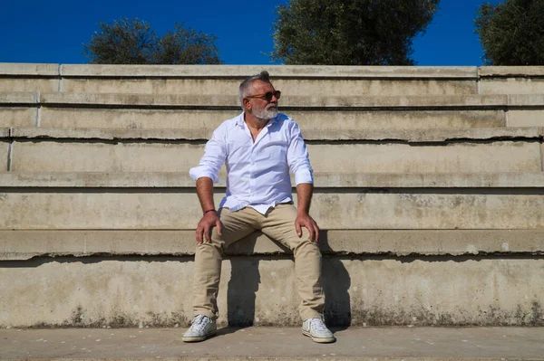Sakallı ve gri saçlı yakışıklı olgun bir adam açık hava konferans salonunun merdivenlerinde oturuyor. Adam farklı yerlere bakıyor. Kıdemli konsept, seyahat ve turizm.
