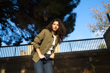 Kıvırcık kahverengi saçlı ve günlük kıyafetler giyen genç ve güzel Latin kadın Avrupa 'da turizm yapıyor, kadın yeşil ceket ve kot pantolon giyiyor. Fotoğraflar için poz veriyor. Turizm konsepti