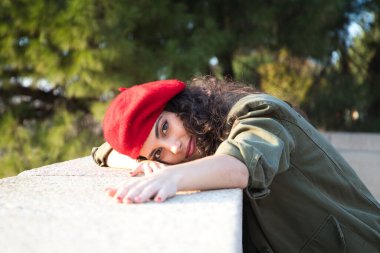 Kıvırcık kahverengi saçlı, kırmızı şapkalı ve günlük kıyafetler giymiş güzel Latin bir kadın Avrupa 'yı geziyor. Fotoğraflar için poz veriyor. Arka planda Akdeniz çam ağacı. Turizm.