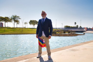 Olgun eşcinsel erkek, yönetici, kır saçlı, sakallı, güneş gözlüklü, ceketli ve kravatlı, sağ bacağında yeni gey gururu bayrağı var. Olgun gay adam kavramı, lgtb gurur, eşitlik, haklar.