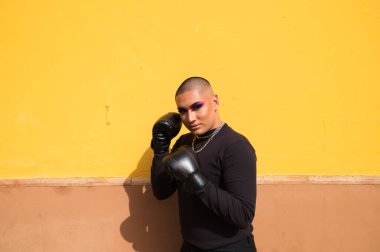 İkili oynamayan genç kişi hardal sarısı arka planda boks eldiveni giyiyor. Makyaj yapıyor ve farklı boks pozları veriyor. Eşitlik kavramı, homoseksüellik, gay, lezbiyen, gay gururu.