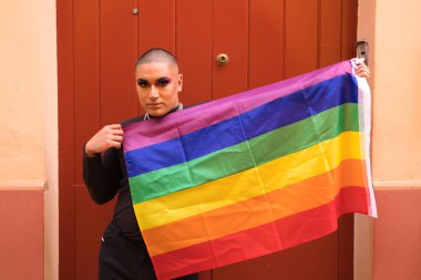 İkili olmayan, Güney Amerika kökenli ve genç, makyajlı ve kırmızı arka planda eşcinsel gurur bayrağı taşıyor. Eşitlik kavramı, homoseksüellik, gay, lezbiyen, gay gururu.