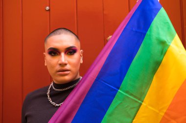 İkili olmayan, Güney Amerika kökenli ve genç, makyajlı ve kırmızı arka planda eşcinsel gurur bayrağı taşıyor. Eşitlik kavramı, homoseksüellik, gay, lezbiyen, gay gururu.