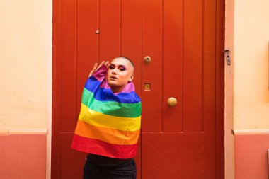 İkili olmayan, Güney Amerika kökenli ve genç biri, makyaj yapıyor ve omuzlarında eşcinsel gururu bayrağı taşıyor. Eşitlik kavramı, homoseksüellik, gay, lezbiyen, gay gururu.