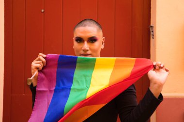 İkili olmayan, Güney Amerika kökenli ve genç, makyajlı ve kırmızı arka planda eşcinsel gurur bayrağı taşıyor. Eşitlik kavramı, homoseksüellik, gay, lezbiyen, gay gururu.