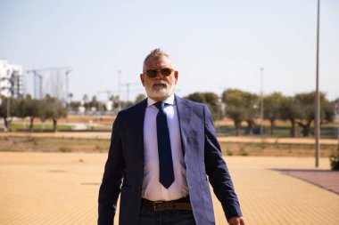 Gri saçlı, sakallı, ceketli, kravatlı ve güneş gözlüklü yakışıklı bir adam işe tekerlekli bavuluyla geliyor. Olgun, idareci adam kavramı.