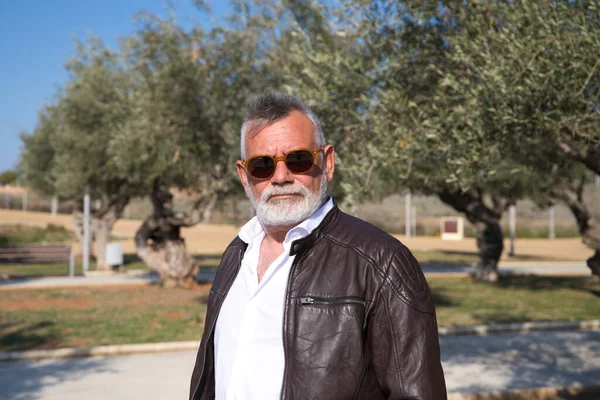 Sakallı ve gri saçlı yakışıklı olgun bir adam parkta yürüyor. Adam bej pantolon, beyaz gömlek ve deri ceket giyiyor. Yaşlılık kavramı