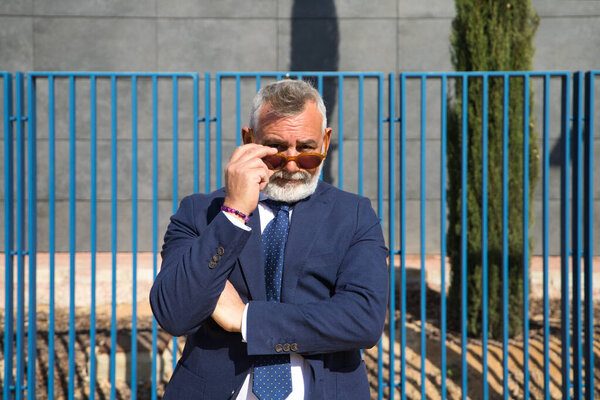 Mature, grey-haired, bearded man with sunglasses, jacket and tie makes different expressions for the photo, looks over his sunglasses and touches his lips. Concept various expressions.