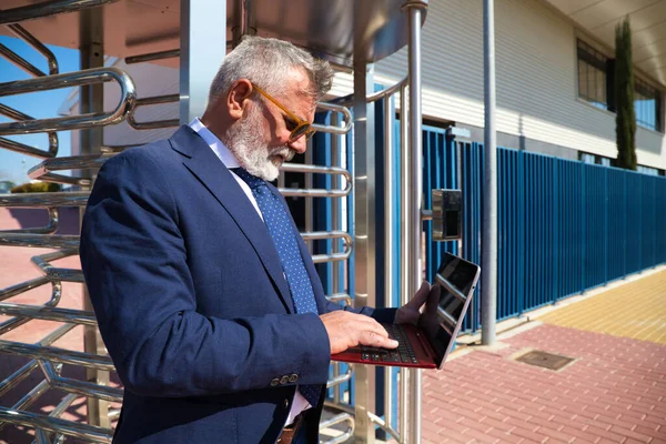 Yetişkin erkek yönetici, gri saçlı, sakallı, güneş gözlüklü, ceketli ve kravatlı sokakta dizüstü bilgisayarına danışıyor. Kavramsal işadamı ve yönetici.