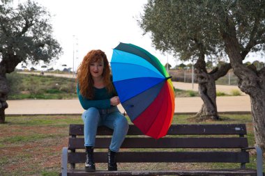 Genç ve güzel kızıl saçlı kadın birçok zeytin ağacının olduğu bir parkta bir bankta oturuyor. Gri günü aydınlatmak için bir renk şemsiyesi var. Moda ve güzellik konsepti