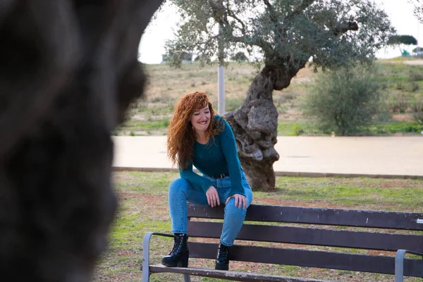 Güzel, genç, kızıl saçlı bir kadın parktaki bir bankta bir sürü zeytin ağacıyla oturuyor. Fotoğrafının çekildiğini fark etmeden gülümsüyor. Moda ve güzellik konsepti
