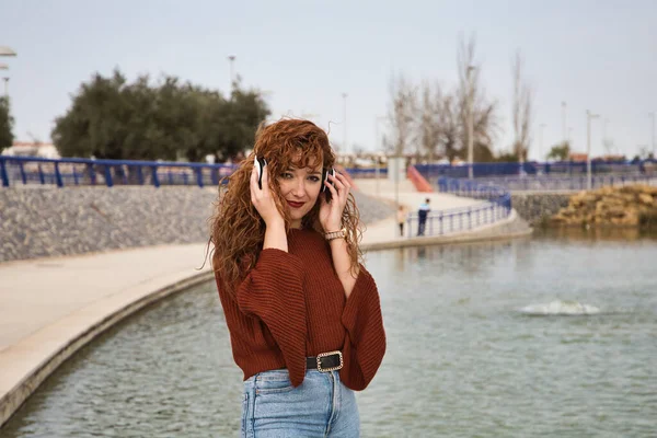 Genç ve güzel kızıl saçlı kadın büyük bir gölün olduğu bir parkta beyaz kulaklıklı müzik dinliyor. Kadın çok mutlu. İfadeler ve mutluluk, müzik ve dans.