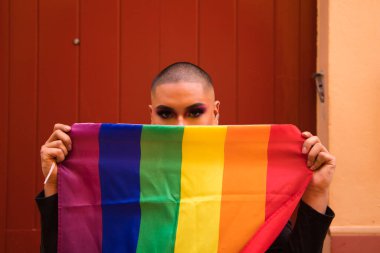 İkili olmayan, Güney Amerika kökenli ve genç, makyajlı ve kırmızı arka planda eşcinsel gurur bayrağı taşıyor. Eşitlik kavramı, homoseksüellik, gay, lezbiyen, gay gururu.