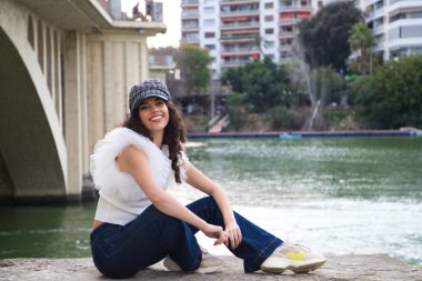 Genç, güzel, siyah, kıvırcık saçlı ve burnu kalkık bir kadın kep takıyor ve nehir kenarında oturuyor. Kadın fotoğraf için poz veriyor. Kavramsal ifadeler. Gülümse, üzül, düşün, eğlen, yaşa.