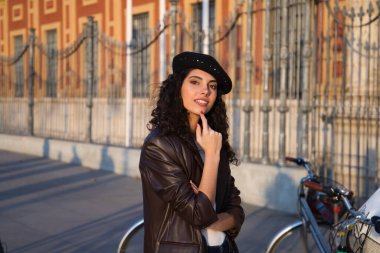 Kameraya doğru kıvırcık saçlı, siyah saçlı genç ve güzel bir kadın. O bir anıtın önünde geziyor ve arka planda bir bisiklet görebilirsiniz. Tatil ve turizm konsepti.