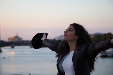 Koyu, kıvırcık saçlı, kollarını havaya açan genç, güzel bir kadın. Şehrin arka planında ve altın saatinde Guadalquivir nehrinde. Turizm ve tatil konsepti.