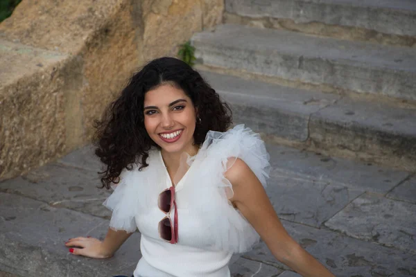 Parktaki bir merdivende oturan siyah, kıvırcık saçlı güzel bir genç kadın. Kadın güneş gözlüğü takıyor ve farklı ifadeler takıyor. İfadeler ve yaşam tarzı konsepti