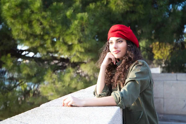 Kıvırcık kahverengi saçlı, kırmızı şapkalı ve günlük kıyafetler giymiş güzel Latin bir kadın Avrupa 'yı geziyor. Fotoğraflar için poz veriyor. Arka planda Akdeniz çam ağacı. Turizm.