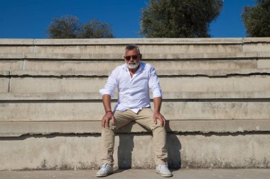 Sakallı ve gri saçlı yakışıklı olgun bir adam açık hava konferans salonunun merdivenlerinde oturuyor. Adam farklı yerlere bakıyor. Kıdemli konsept, seyahat ve turizm.