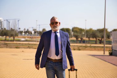 Gri saçlı, sakallı, ceketli, kravatlı ve güneş gözlüklü yakışıklı bir adam işe tekerlekli bavuluyla geliyor. Olgun, idareci adam kavramı.