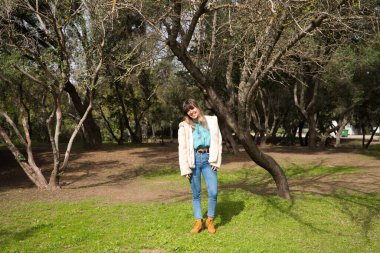 Kot pantolon giymiş, beyaz şapkalı ve botlu genç ve güzel sarışın kadın parkta yürüyor ve ağaçların arasında güneşli bir günün tadını çıkarırken fotoğrafını çekiyor. Boş zaman kavramı.