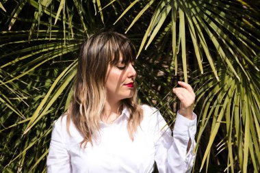 Güzel, sarışın, genç bir kadın parkta yeşil palmiye ağaçlarının önünde farklı ifadeler takınarak eğleniyor. Tatil ve boş zaman kavramı. Çeşitli ifadeler.