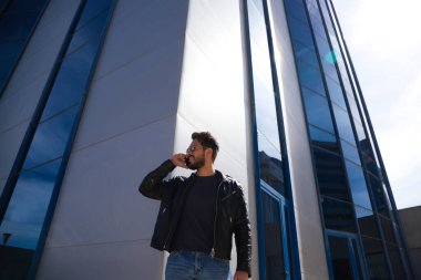 Sakallı, vücudu yontulmuş ve güneş gözlüklü yakışıklı genç adam cam bir binanın önünde telefonla konuşuyor. Adam kot pantolon ve deri ceket giyiyor..