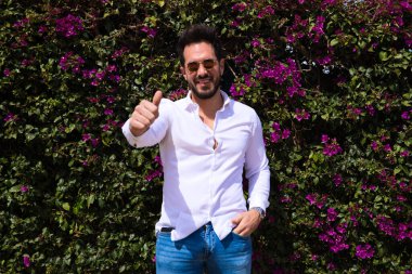 Sakallı, yontulmuş vücutlu yakışıklı bir genç adam portresi. Arka planda yeşil ve mor tırmanma bitkileri var. Adam elini kaldırırken dümdüz ileriye bakıyor..