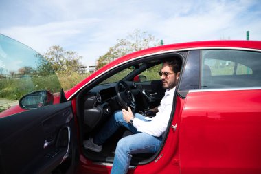 Genç yakışıklı bir adam, kırmızı spor arabasında oturan heykel gibi vücudu var. Adam zengin ve modern kıyafetler giymiş. Yüksek yaşam standardı ve finansal olarak iyi konumlandırılmış..