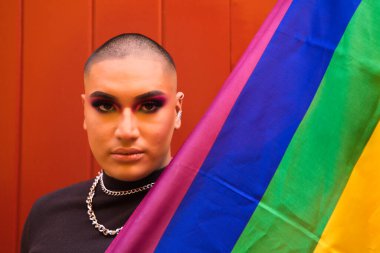 Non-binary person, of South American origin and young, the person is wearing make-up and holding the gay pride flag on a red background. Concept equality, homosexuality, gay, lesbian, gay pride.