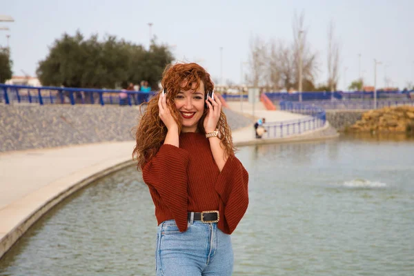 Genç ve güzel kızıl saçlı kadın büyük bir gölün olduğu bir parkta beyaz kulaklıklı müzik dinliyor. Kadın çok mutlu. İfadeler ve mutluluk, müzik ve dans.