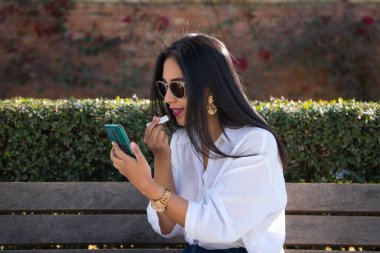 Avrupa 'da tatilde olan genç ve güzel bir Güney Amerikalı kadın cep telefonunu ayna olarak kullanarak bir bankta dudaklarını boyuyor..