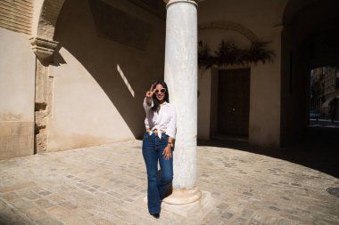 Genç, güzel ve esmer Güney Amerikalı kadın Avrupa 'da seyahat ediyor ve parmaklarıyla zafer işareti yaparken beyaz mermer bir sütuna yaslanıyor. Seyahat ve tatil konsepti.