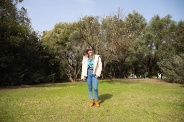 Kot pantolon giymiş, beyaz şapkalı ve botlu genç ve güzel sarışın kadın parkta yürüyor ve ağaçların arasında güneşli bir günün tadını çıkarırken fotoğrafını çekiyor. Boş zaman kavramı.