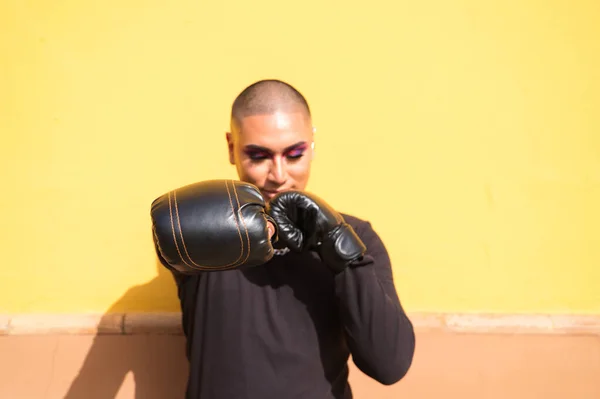 Young non-binary person wears boxing gloves on a mustard yellow background, the person is make up and does different boxing poses. Concept equality, homosexuality, gay, lesbian, gay pride.