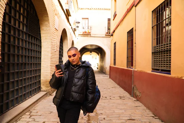 Non-binary and young person from South America is on holiday in Europe, the person is make up and wearing black clothes. He is walking around the city consulting his mobile phone. Concept of equality.