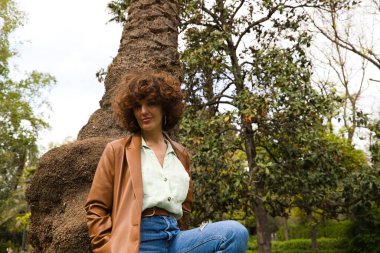 Kıvırcık saçlı güzel İspanyol kadın parkta bir ağaç gövdesine yaslanıyor. Kadın mutlu ve şehirde tatilinin tadını çıkarıyor. Mutluluk, moda ve güzellik kavramı.