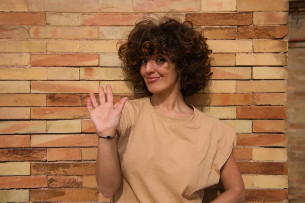 Beautiful, happy woman waving with palm of hand. The woman has a brick wall in the background. Greetings, hello, goodbye, welcome, see you later, see you.
