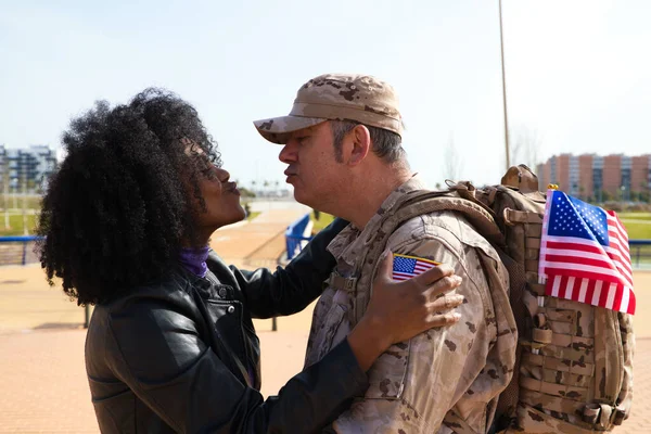 Beautiful Afro-American woman and American soldier kiss. The soldier has come from war and special missions. The woman\'s hand is seen next to the US flag. Concept of war and army.