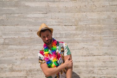 Havai çiçekli gömlek, şapka ve renkli çiçekli kolye giyen yakışıklı bir genç. Adam çok mutlu. Arka plan gri duvar. Seyahat ve tatil konsepti.