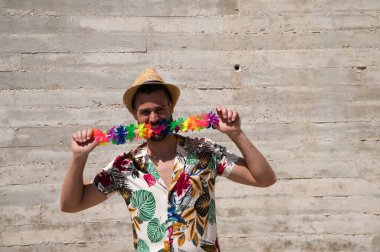 Havai çiçekli gömlek, şapka ve renkli çiçekli kolye giyen yakışıklı bir genç. Adam yüz ve vücut ifadeleriyle farklı jestler yapıyor. Arkaplan gri duvarı.