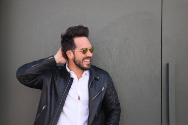 Sakallı, vücudu yontulmuş ve güneş gözlüklü yakışıklı genç adam portre için farklı pozlar verirken garajının gri kapısında duruyor. Farklı ifadeleri kavra.