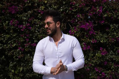 Sakallı, yontulmuş vücutlu yakışıklı bir genç adam portresi. Arka planda yeşil ve mor tırmanma bitkileri var. Fotoğraflar için farklı yerlere bakıyor..