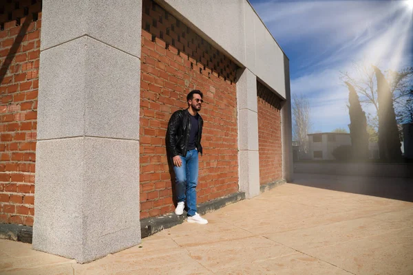 Orta yaşlı, yakışıklı ve esmer, sakallı ve tuğla bir duvarda heykeli olan bir adam. Siyah deri ceketli, aynalı güneş gözlüklü ve kot pantolonlu bir adam. Adam fotoğraf için poz veriyor. Erkek moda konsepti.