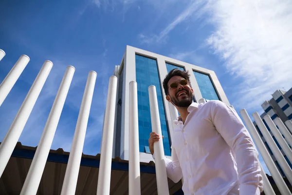 Sakallı, vücudu yontulmuş ve güneş gözlüklü yakışıklı genç adam beyaz çitlere tutunuyor. Fotoğraf aşağıdan çekildi ve arka planda mavi gökyüzünü görebilirsiniz..