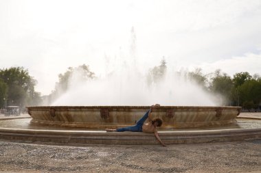 Dansçı olan genç ve güzel bir kadın bir meydanda bacakları açık bir çeşmede duruyor. Kadın atletik ve esnektir. Refah ve sağlık konsepti.